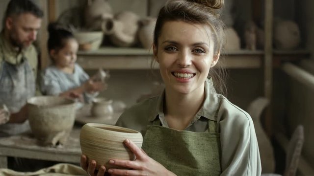 PAN of happy female artisan holding ceramic bowl and smiling while posing for camera in pottery workshop; man and children sculpting with clay in background