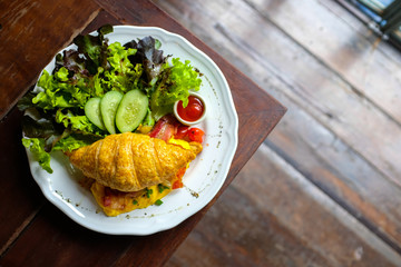 Flat lay of croissant and salad served in white plate on wooden table with blurred wooden floor background in cafe