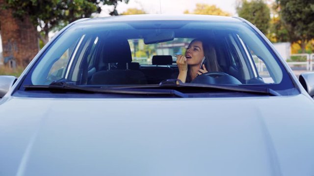 A happy young woman doing makeup in the car looking in the mirror and at the same time talking on the phone with friends. 4K UHD video.