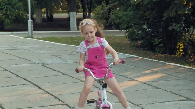 A happy, beautiful, little girl with long blond hair in a pink skirt and jumper rides a children's bike on the road, she smiles. Super slow motion