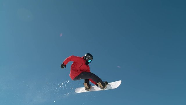 SLOW MOTION CLOSE UP: Extreme snowboarder jumping across the clear blue sky in sunny mountain wilderness. Freeride boarder snowboarding and flying over camera in mountain ski resort. Snowboard jump