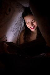 young woman reading a book under the covers and smile