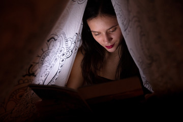 young woman reading a book under the covers