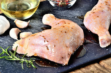 Close-up of raw spicy chicken legs ready for baking on a cutting board with a fresh rosemary, garlic, cumin, jug with oil, pepper and salt on a wooden table