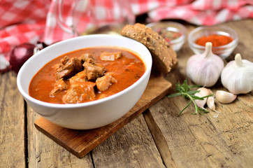 Pork goulash with pieces of meat in a bowl, garlic, pepper, onion, jug with oil and red checkered...