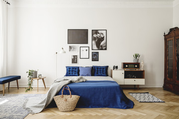 Navy blue bed with blanket next to cabinet with radio in retro bedroom interior with posters. Real photo