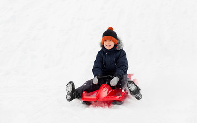 childhood, sledging and season concept - happy little boy sliding on sled down snow hill outdoors in winter