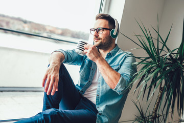 Photo in of pleased man looking through window and listening to music using wireless earphones while drinking cup of coffee