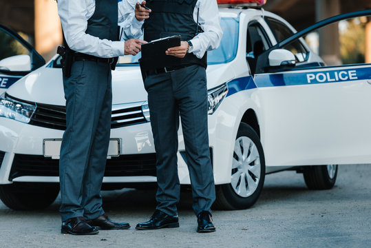 partial view of male police officer with clipboard and pointing by finger to colleague standing near car at street - Powered by Adobe