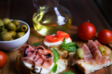 Appetizing bruschettes with jamon, tomatoes, basil, mozzarella, olives and oil on a dark wooden board