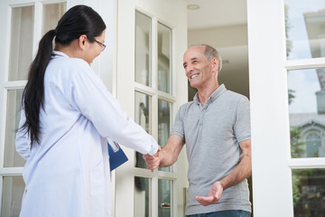Female doctor with clipboard shaking hand of senior man during home visit while entering house