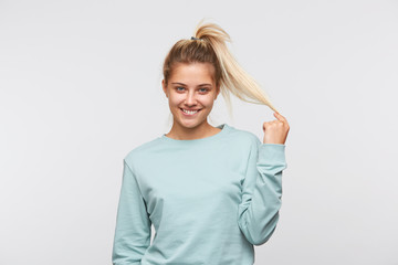Portrait of smiling playful young woman with blonde hair and ponytail wears blue sweatshirt biting...