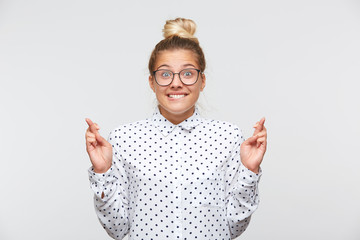 Portrait of happy excited young woman with bun wears polka dot shirt and spectacles keeps fingers...