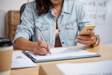 Young woman sitting at the table and making notes in document while holding mobile phone
