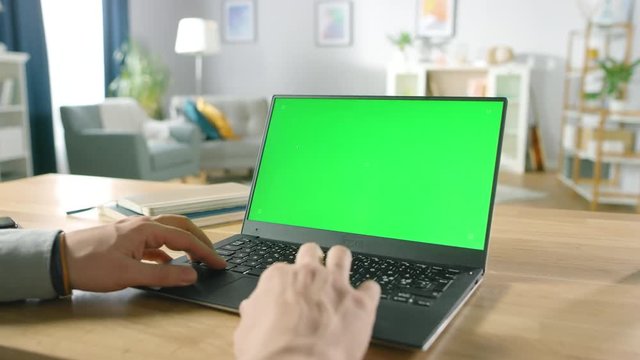 Close-up of a Man Uses Laptop with Green Mock-up Screen While Sitting at the Desk in His Cozy Living Room.