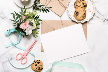 Marble desk with bouquet of flowers, pink scissors, postcard, kraft envelope, cotton branch, oat cookies, invitation card with copy space for text. Flat lay, top view. Woman's day or mother's day.