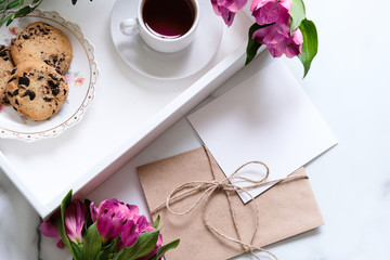 Marble desk with cup of coffee, pink flowers, postcard, kraft envelope, twine, cotton branch, oat cookies, invitation card with copy space for text. Flat lay, top view. Woman's day or mother's day.