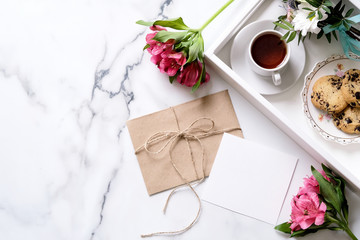 Marble desk with cup of coffee, pink flowers, postcard, kraft envelope, twine, cotton branch, oat cookies, invitation card with copy space for text. Flat lay, top view. Woman's day or mother's day.