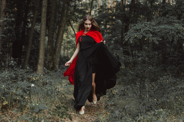 elegant mystic girl in black dress and red cloak running in woods