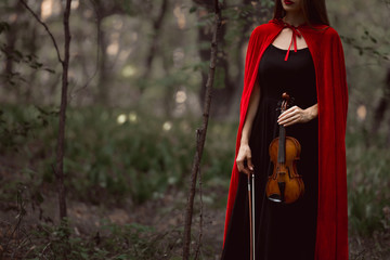 cropped view of elegant woman in red cloak holding violin in dark woods