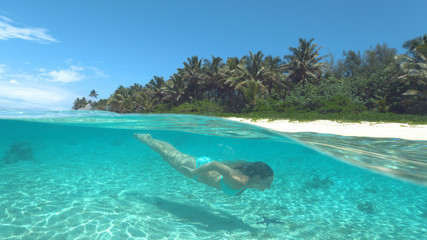 HALF UNDERWATER: Happy young woman in bikini diving near sunny tropical island.