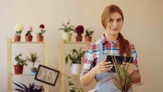 Small business. A girl in a striped apron and a plaid shirt, standing in her shop and holding a digital tablet, is planning a bouquet. A young girl looks up from the tablet and smiles.