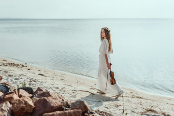 beautiful elegant woman in white dress holding violin and walking on beach near sea © LIGHTFIELD STUDIOS