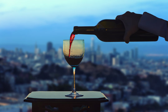 Female hand with bottle pours red wine into glasses on blurred San Francisco city background. Service on the roof of the restaurant