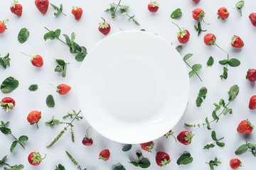 top view of empty plate surrounded with ripe strawberries and mint leaves on white tabletop