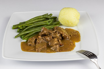 Fleischgericht als Goulasch auf einem Teller mit Beilagen wie Bohnen und Kartoffelkloß