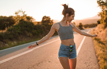 Young girl with hair bun and denim shorts dancing walking and smiling on the edge of the roadway in a sunset of summer