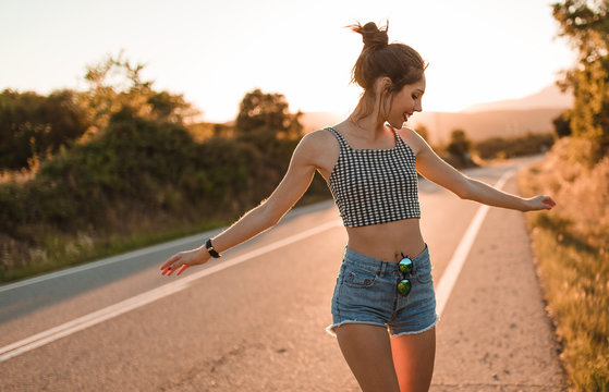 Young girl with hair bun and denim shorts dancing walking and smiling on the edge of the roadway in a sunset of summer