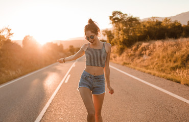Young girl with hair bun and denim shorts dancing walking and smiling on the edge of the roadway in a sunset of summer