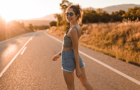 Young girl with hair bun and denim shorts dancing walking and smiling on the edge of the roadway in a sunset of summer