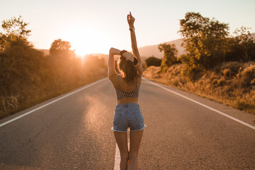 Young girl with hair bun and denim shorts dancing walking and smiling on the edge of the roadway in a sunset of summer