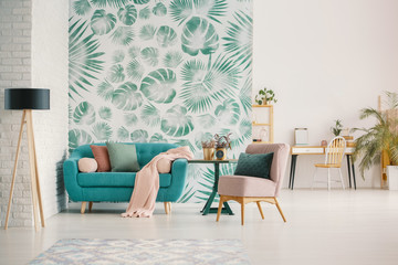 Pink chair and blue sofa in spacious living room interior with leaves wallpaper and lamp. Real photo
