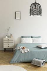 A double bed with sage green and white bedding standing on a wooden floor in a bright bedroom interior. A nightstand next to the bed. Real photo