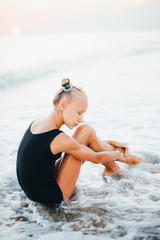 A teenage girl is sitting on the beach in a black swimsuit.