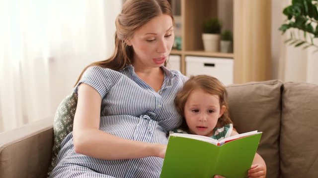pregnancy, people and family concept - happy pregnant mother and little daughter reading book at home