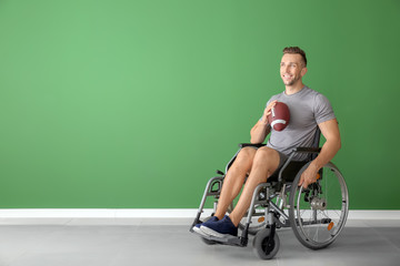 Fototapeta na wymiar Young man with rugby ball sitting in wheelchair against color wall