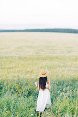 Girl walking on the field, in a hat and summer dress. Smiling and laughing, beautiful sunset in the forest and in nature. Happy traveler, lifestyle.