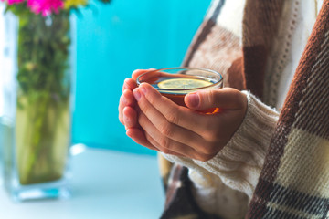 Obraz premium Tea drinking. The woman in a knitted, white pullover and in a warm plaid holds a cup of hot tea with lemon in her hands. Cozy weekends, winter drinks. Ceylon, Chinese tea