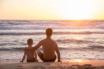 Father shows sunset on the sea to his son sitting on the sand