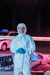male criminologist in protective suit and mask looking at camera near crime scene with corpse in body bag and case with investigation tools