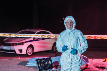 mature male criminologist in protective suit and mask looking at camera near crime scene with corpse in body bag