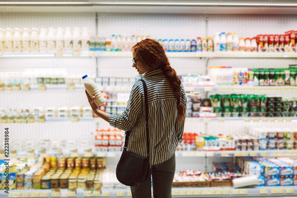 Wall mural Pretty girl in eyeglasses and striped shirt choosing milk in dairy department in supermarket