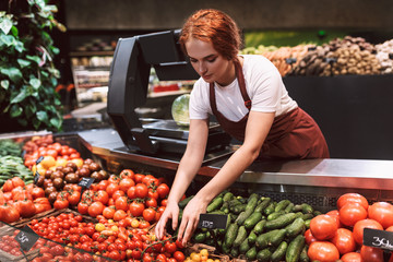 Young seller in apron standing behind counter with vegetables thoughtfully working in supermarket
