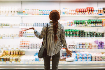 Young woman in striped shirt from back trying to choose dairy products in modern supermarket