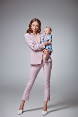full length view of stylish young mother holding adorable infant daughter and looking at camera on...