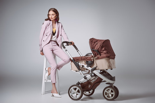 fashionable young woman with baby carriage sitting on stool and looking away on grey